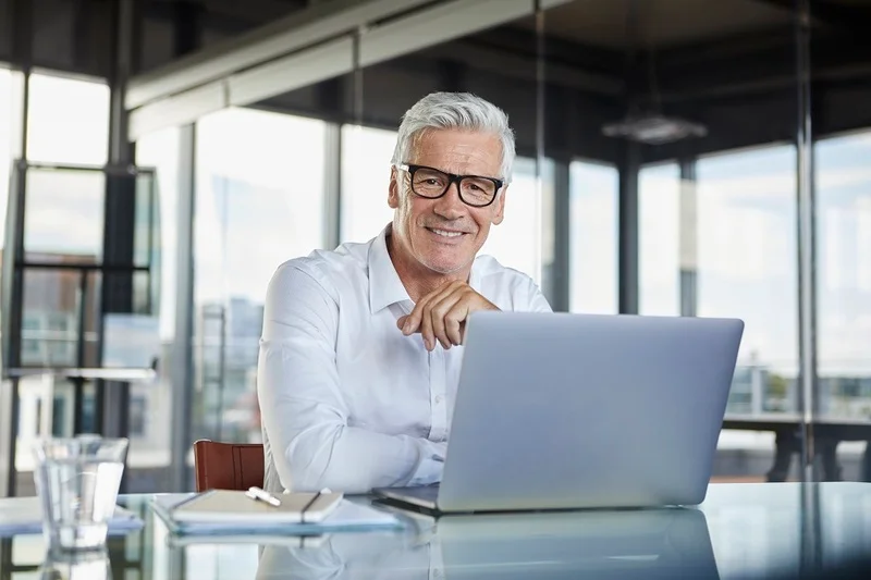 A Smiling Professional Working On An Open Laptop, Illustrating How To Add Resume To Linkedin.